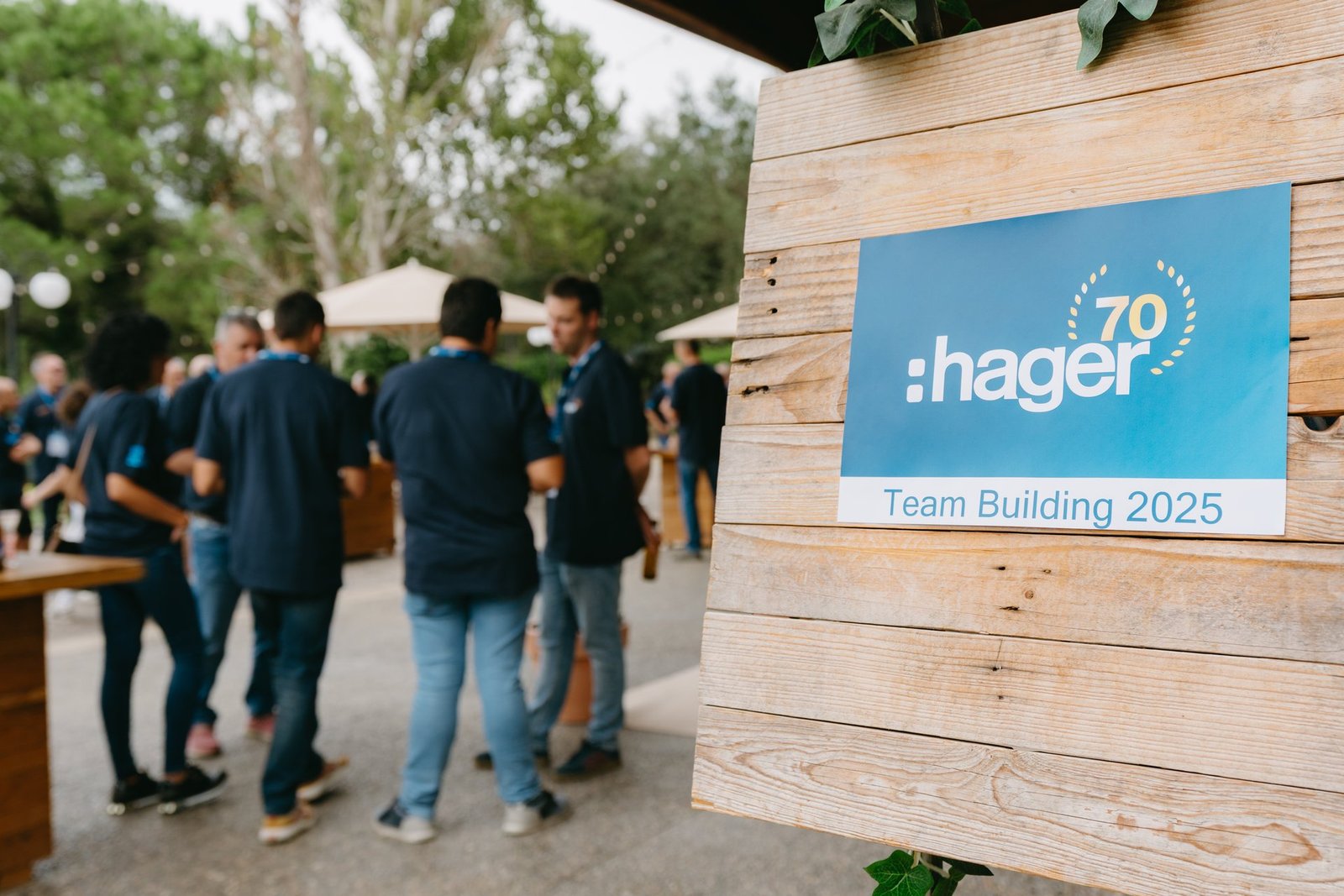 Cartel de madera con logo Hager 70 aniversario y 'Team Building 2025'. Empleados conversando en evento al aire libre.